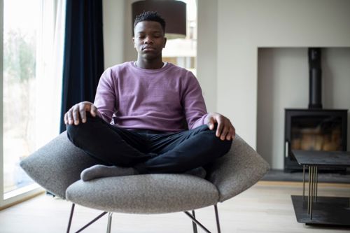 Male teenager in a purple jumper sitting on a grey armchair with crossed legs and eyes closed.