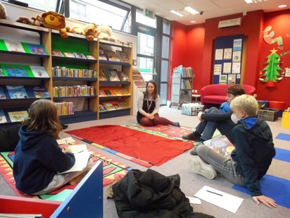 Group of students sat on the floor in a red classroom being spoken to by afemale teacher wearing a lanyard