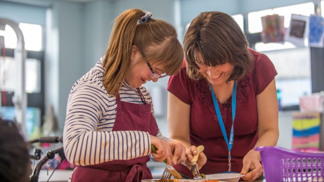 Learner with Downs Syndrome in a stripy top and apron laughing with a teacher while they cut up some food.