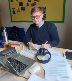 Caucasian teenage male with blonde hair and glasses smiling at a laptop while writing on paper